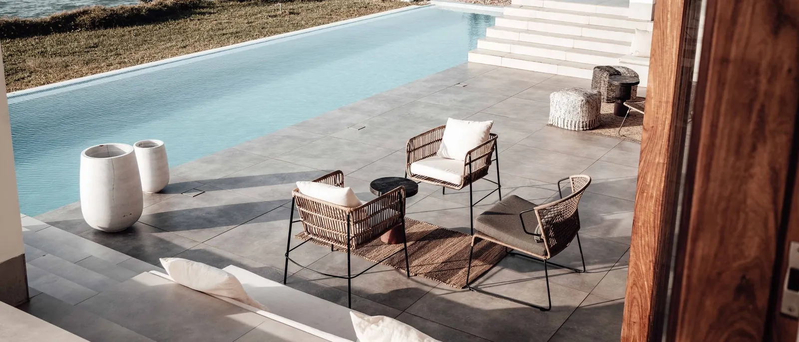 Terraza de una vivienda moderna con piscina, sillones de ratán y escalera blanca, junto al mar bajo un sol claro