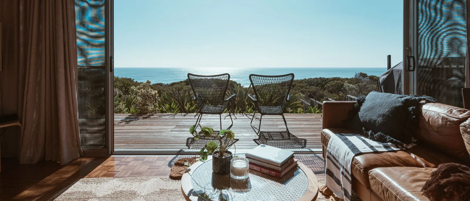 Salón con la puerta corredera abierta a una terraza de madera con dos sillas y vistas al mar
