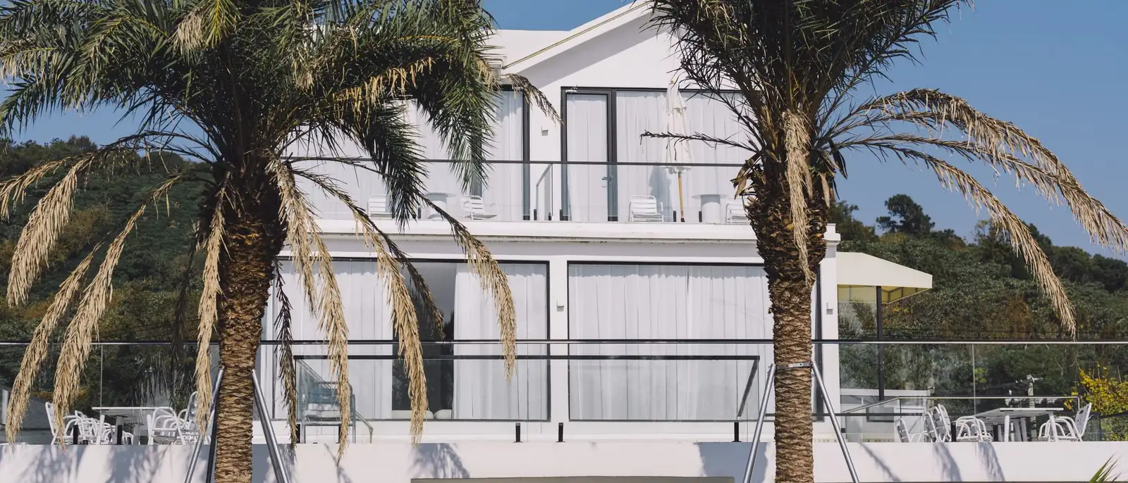 Casa moderna de paredes blancas con dos plantas y terrazas con barandilla de cristal, entre dos palmeras bajo un cielo azul