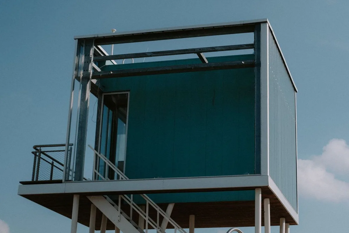 Módulo de vivienda verde azulado elevado sobre pilares blancos, con escalera, balcón y puerta acristalada, bajo un cielo azul