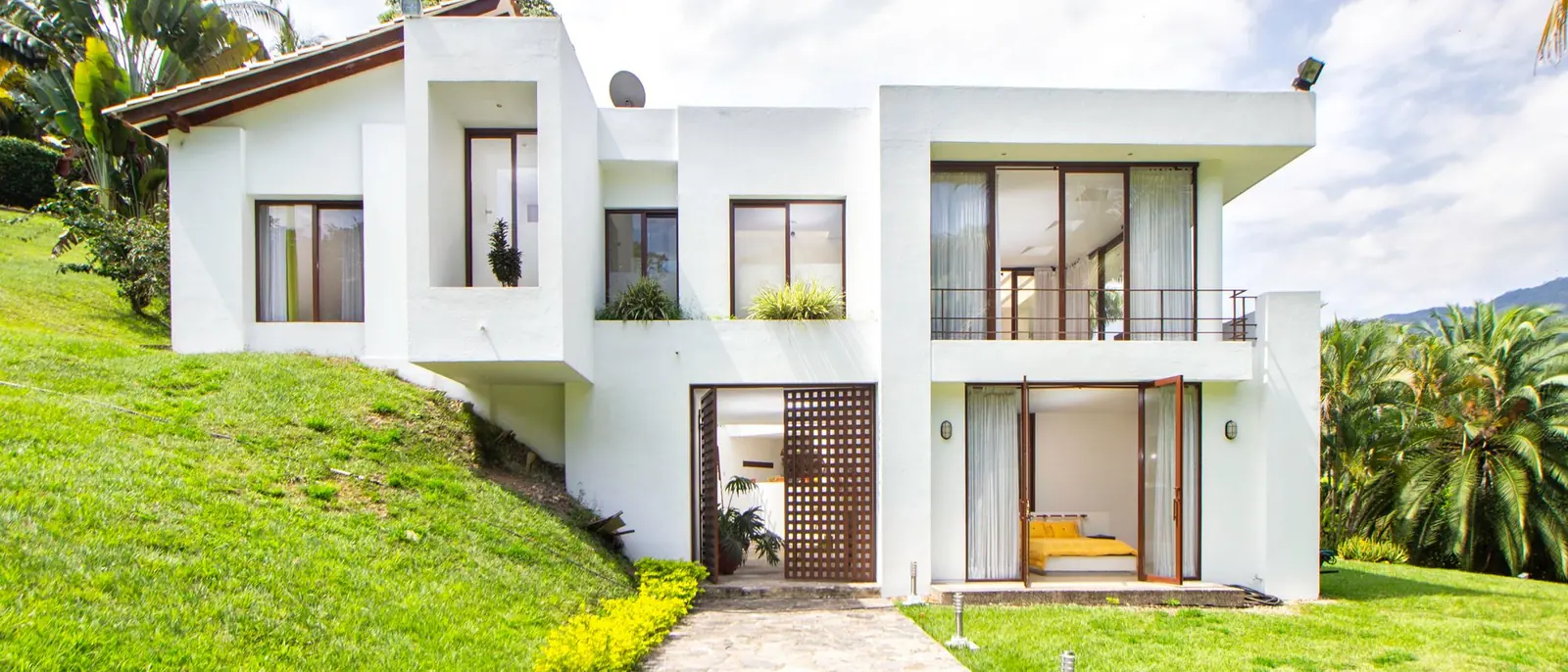Vivienda moderna de muros blancos en una ladera de césped, con palmeras alrededor y nubes en el cielo
