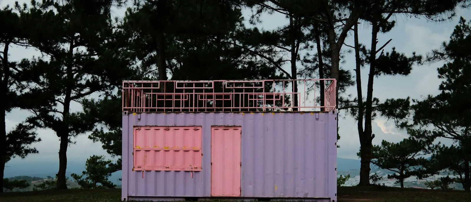 Contenedor marítimo lila convertido en caseta, con una puerta y una contraventana rosas y una barandilla rosa en el techo, sobre un claro de hierba y tierra entre pinos