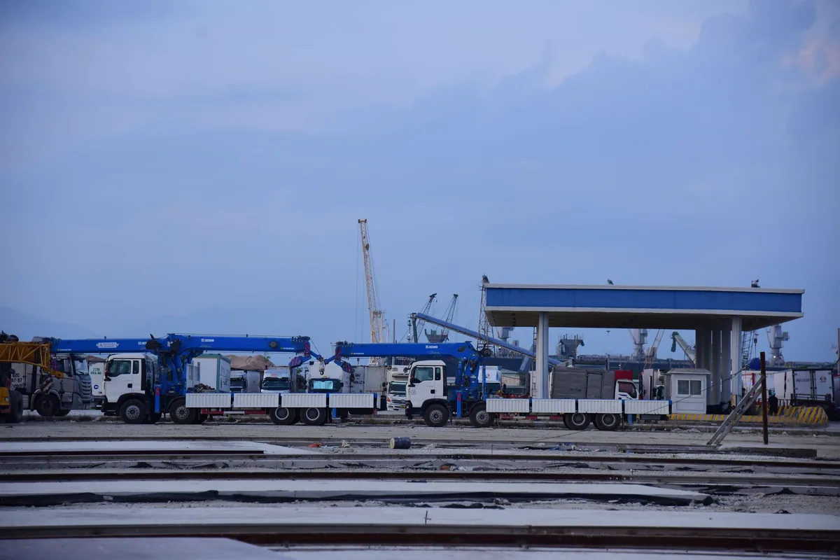 Camiones con grúa articulada aparcados en una zona de carga, con grúas de puerto al fondo