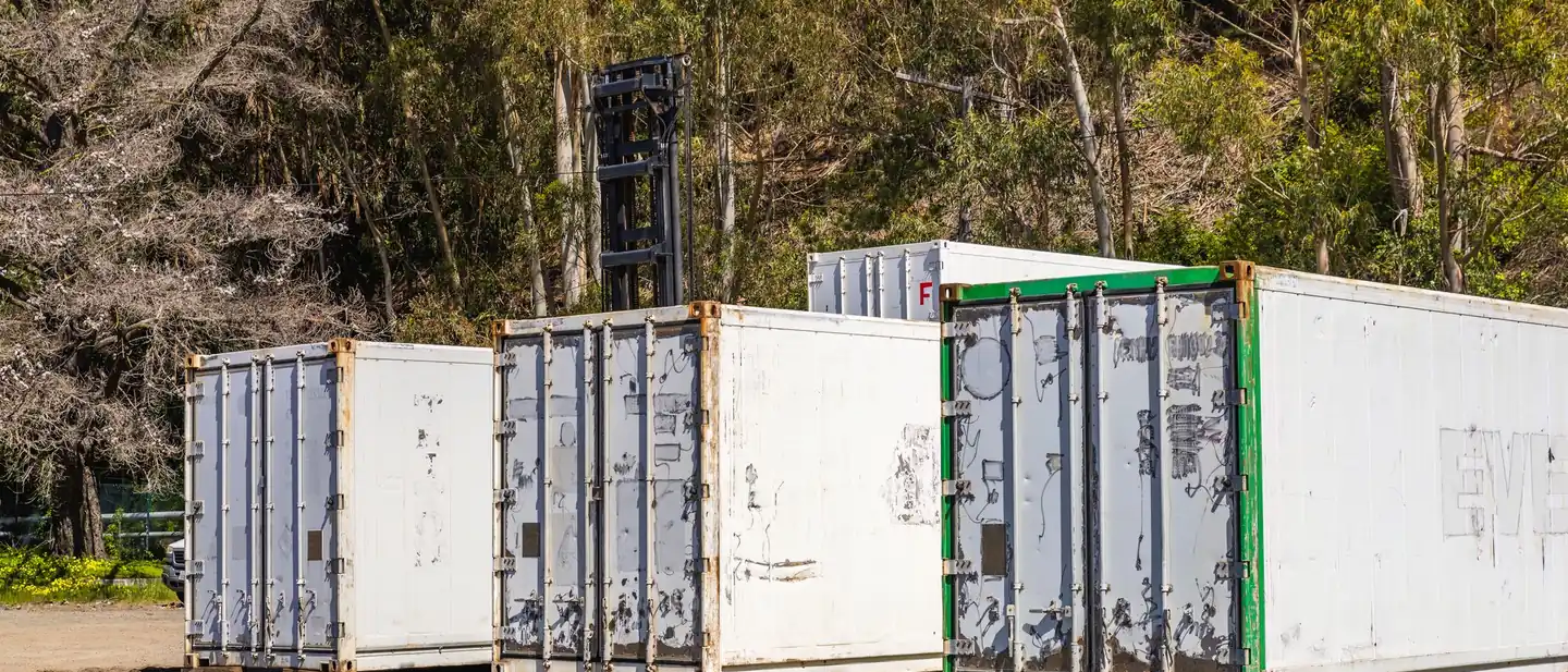 Contenedores marítimos blancos apoyados sobre un terreno de grava junto a un arbolado