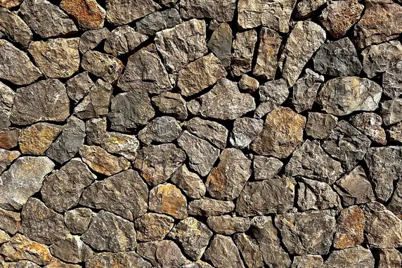 Muro de piedra seca hecho con piedras oscuras e irregulares, con tonos grises y ocres, iluminado por el sol